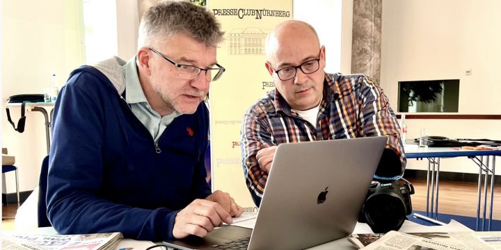 Zwei Männer sitzen an einem Laptop-Bildschirm, auf dem Schreibtisch liegen einige Zeitungen